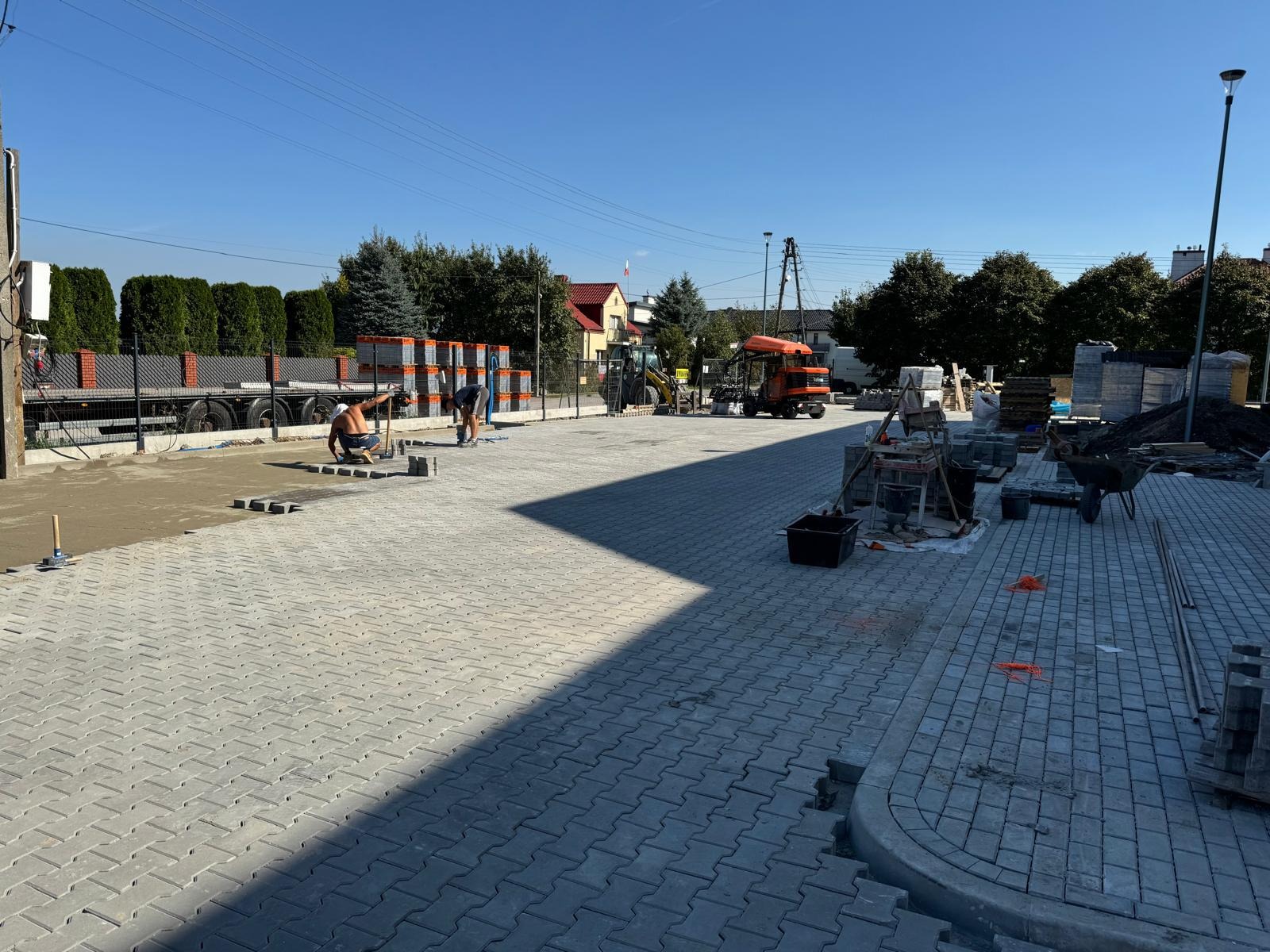 Pracownicy układają szarą kostkę brukową na placu, widoczne maszyny budowlane i stosy kostki w tle, silne oświetlenie słoneczne tworzy wyraźny cień na powierzchni.