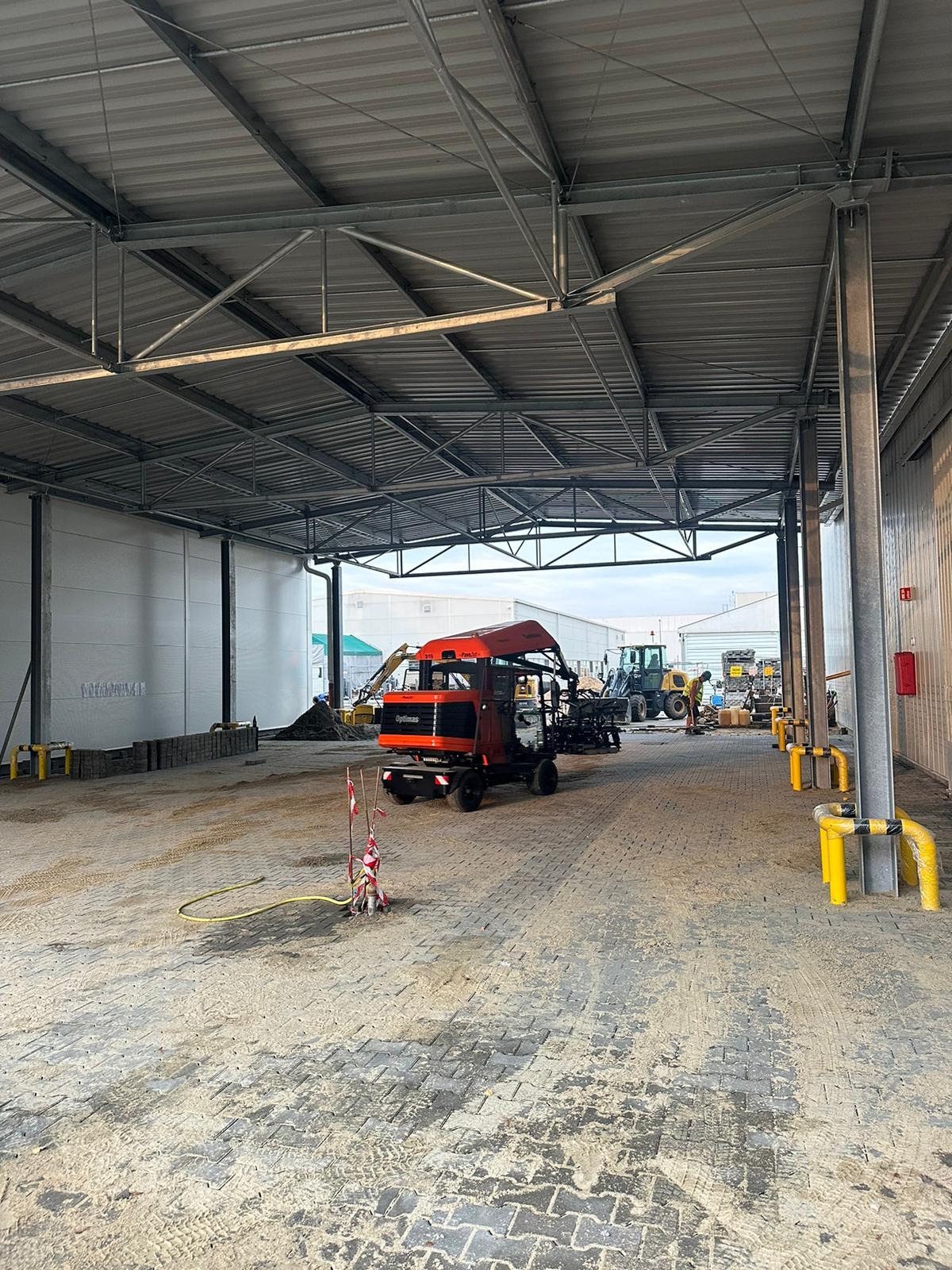 Pomarańczowy miniładowarka Optimas na placu budowy z częściowo ułożoną kostką brukową, widoczne inne maszyny budowlane w tle, hala z metalową konstrukcją dachu.