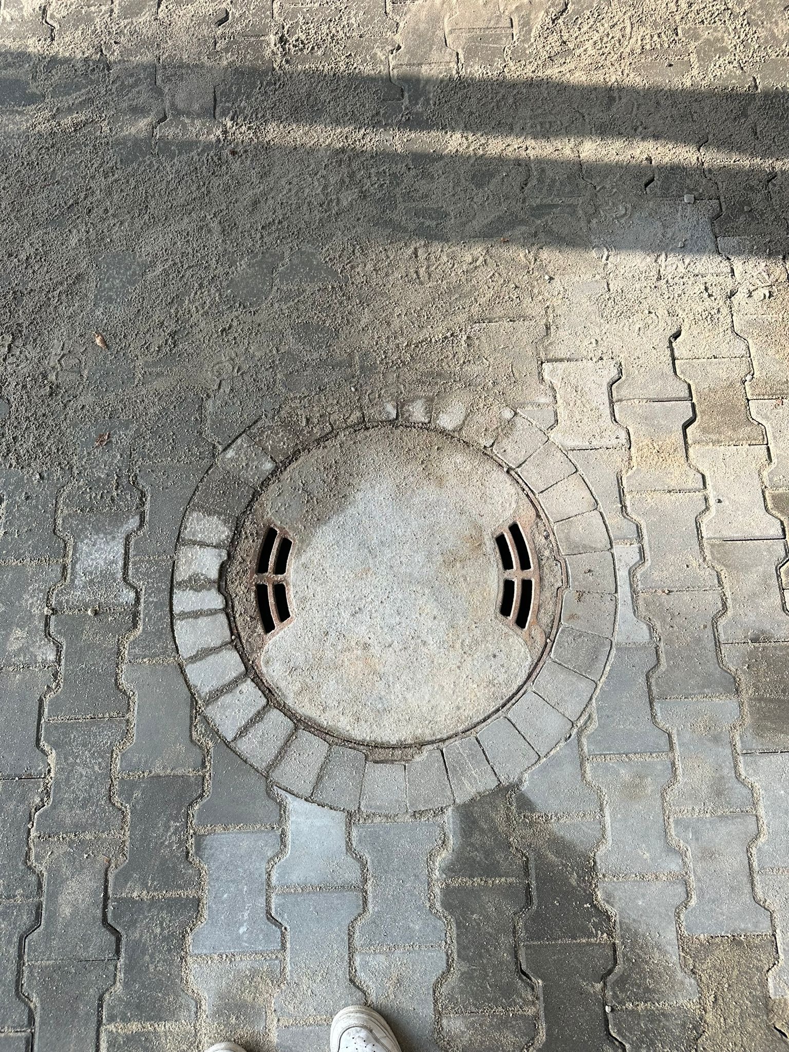 Szara, betonowa studzienka kanalizacyjna z czterema otworami, otoczona kostką brukową ułożoną w okrąg, z widocznymi śladami piasku i ziemi na powierzchni.