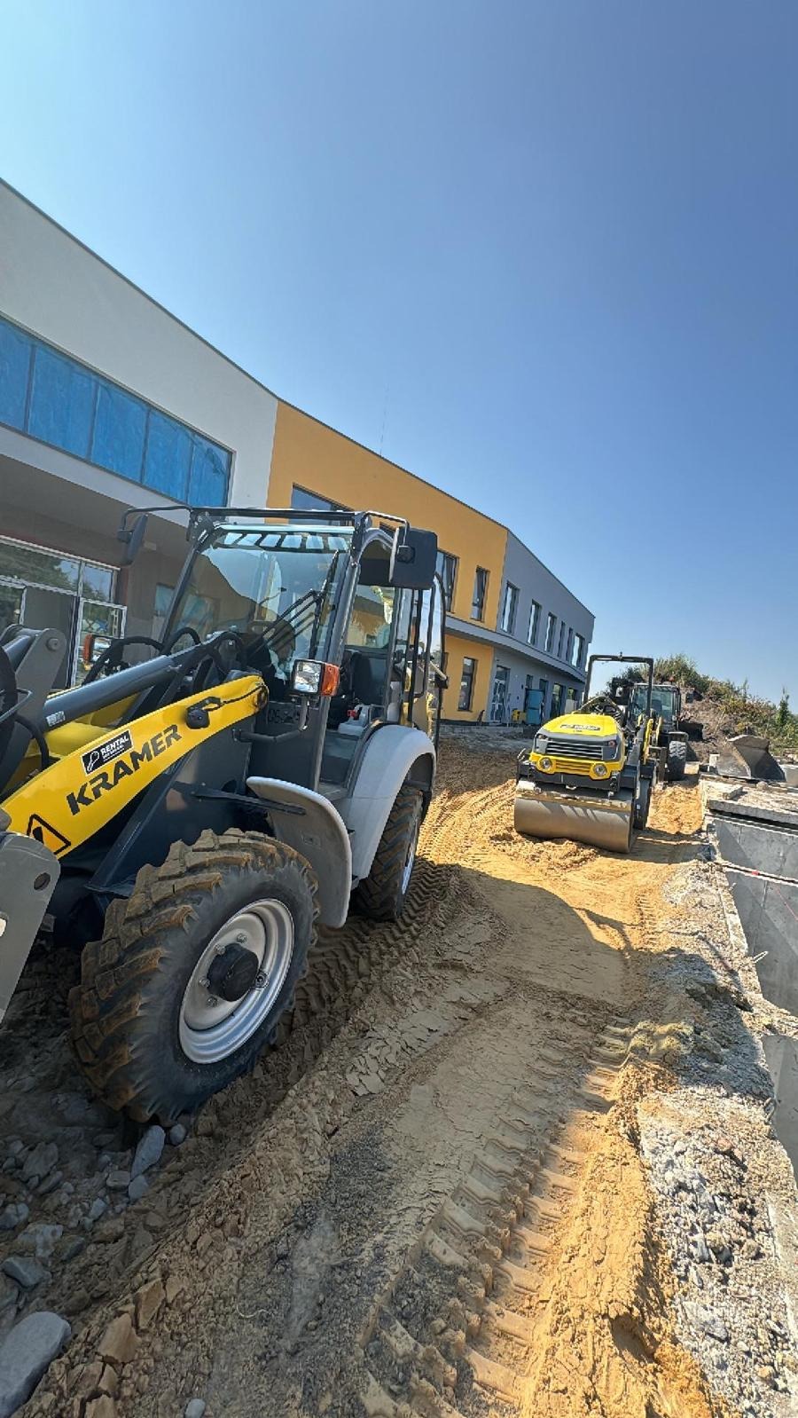 Żółto-szara ładowarka Kramer i walec drogowy na placu budowy z częściowo wyrównanym terenem, widoczne ślady opon na piasku i fragment budynku w tle.