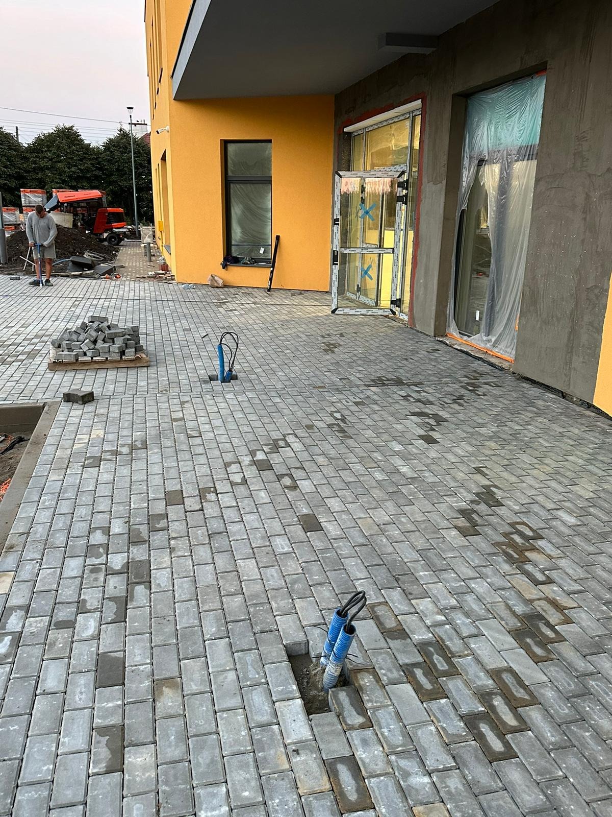 Szara kostka brukowa ułożona przed budynkiem z żółtą elewacją, widoczne otwarte drzwi i okna w folii ochronnej, w tle robotnik z taczką i wykopane miejsce na kable.