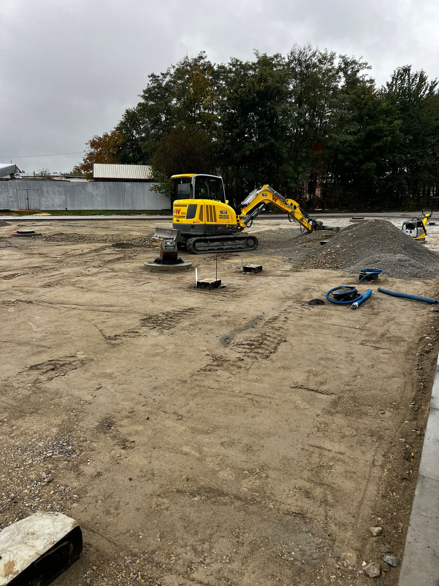 Żółta minikoparka Wacker Neuson na placu budowy, w tle hałda gruzu i ubijarka, ślady opon na ziemi.