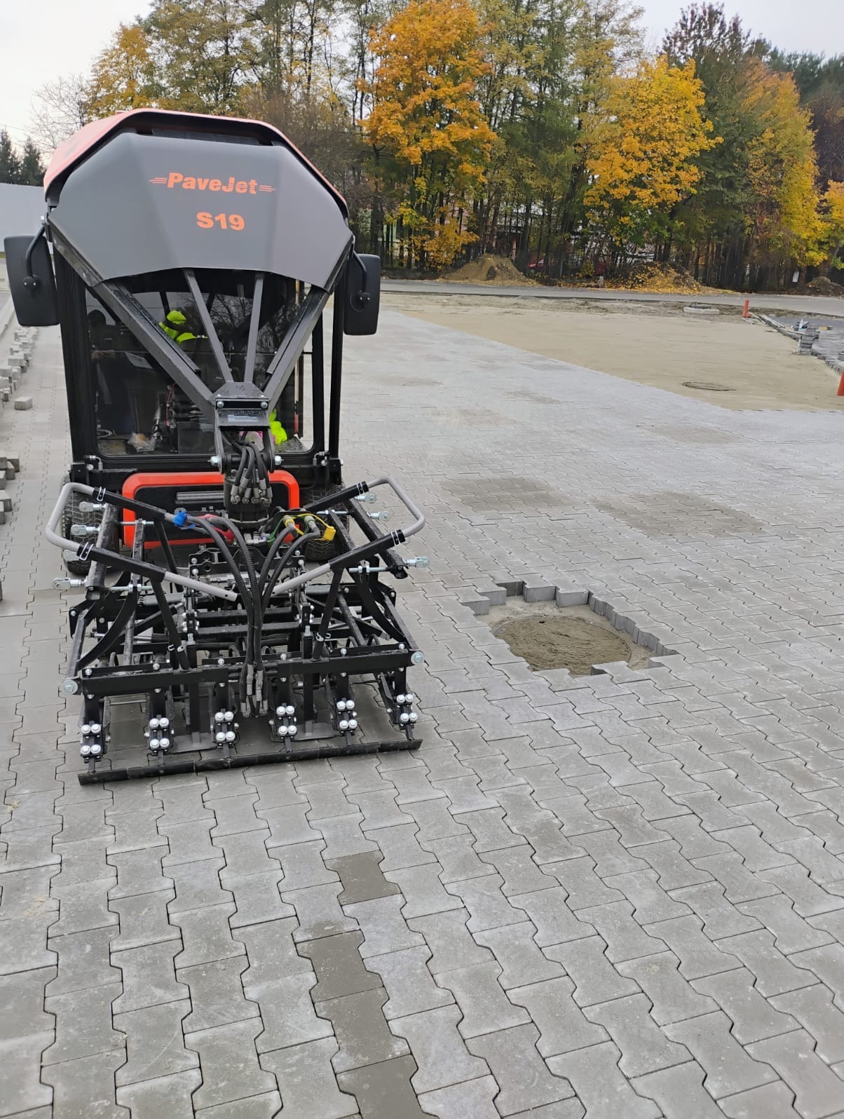 Maszyna PaveJet S19 układająca szarą kostkę brukową typu 'I'. Widoczny fragment terenu przygotowany pod kostkę oraz operator maszyny w odblaskowej kamizelce. W tle drzewa z jesiennymi liśćmi.