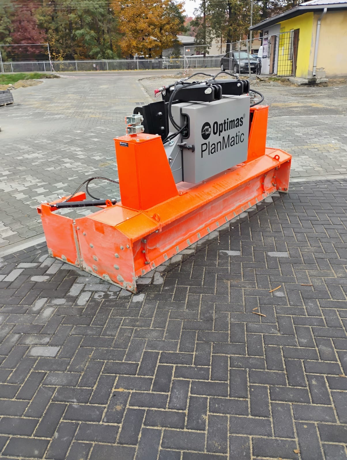 Maszyna Optimas PlanMatic, pomarańczowo-szara, do precyzyjnego wyrównywania terenu pod kostkę brukową, na tle ułożonej kostki w jodełkę, widoczny budynek w tle.