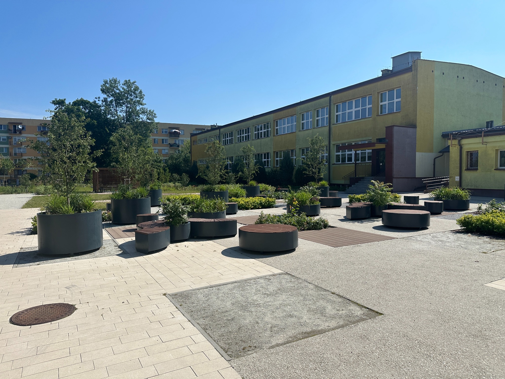 Ułożona kostka brukowa w przestrzeni publicznej z donicami z roślinami i siedziskami, na tle budynku szkolnego i bloku mieszkalnego w słoneczny dzień.