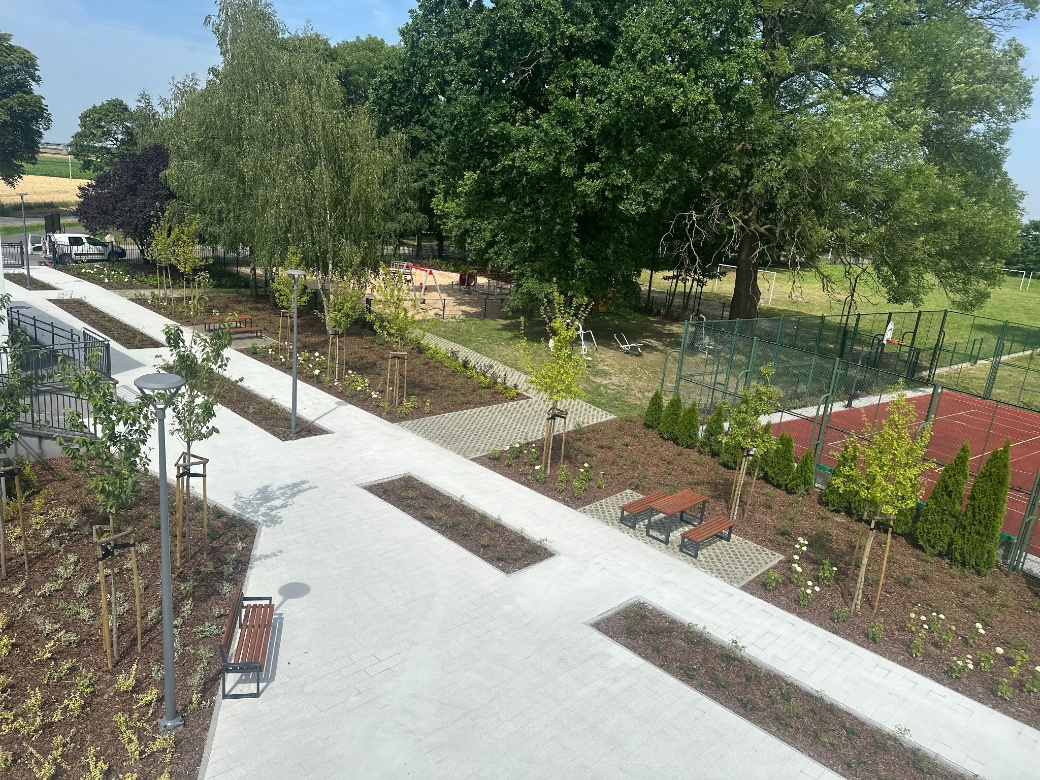 Nowo wyłożony chodnik z jasnoszarej kostki brukowej, z wkomponowanymi rabatami ziemnymi i nasadzeniami, ławkami parkowymi oraz młodymi drzewkami, w tle plac zabaw i boisko sportowe.