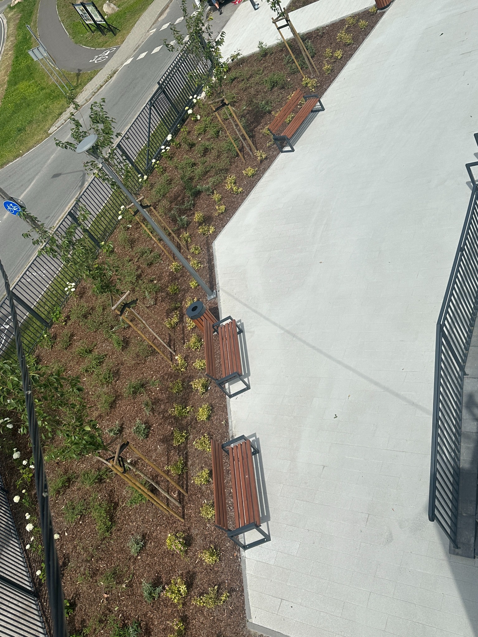 Widok z góry na nowo zagospodarowaną przestrzeń miejską z elementami zieleni, drewnianymi ławkami i jasną nawierzchnią chodnika, otoczoną metalowym ogrodzeniem i fragmentem ulicy.