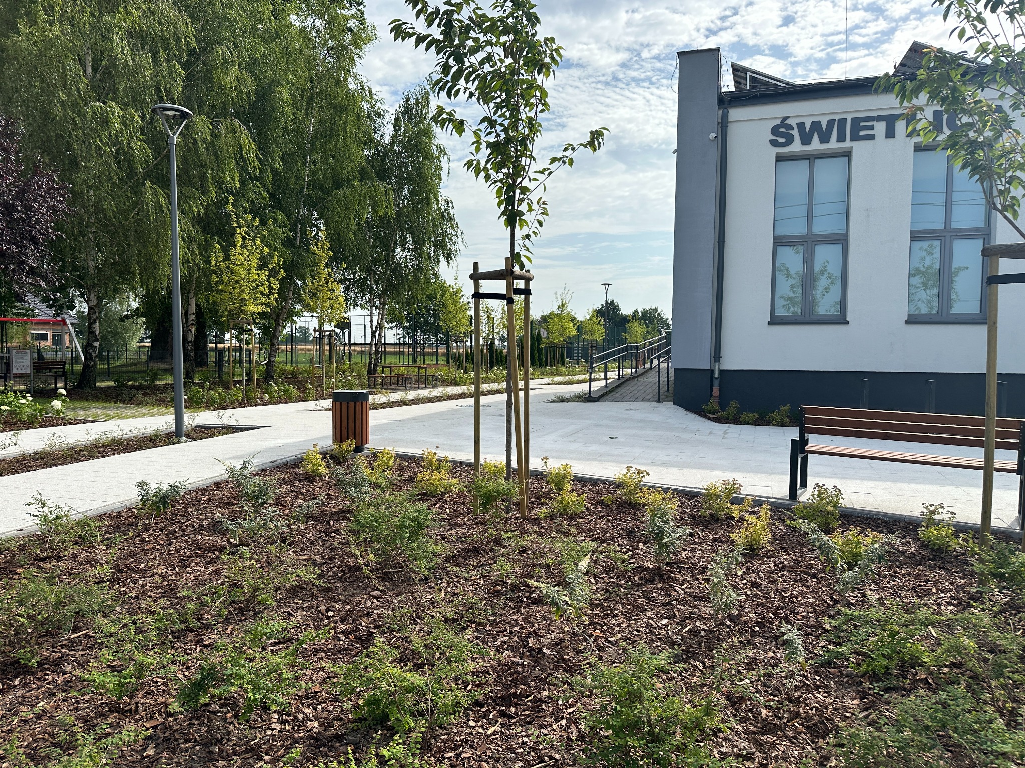 Świeżo założony rabat kwiatowy z młodymi drzewkami podpartymi palikami, obok chodnika z kostki i budynku użyteczności publicznej.