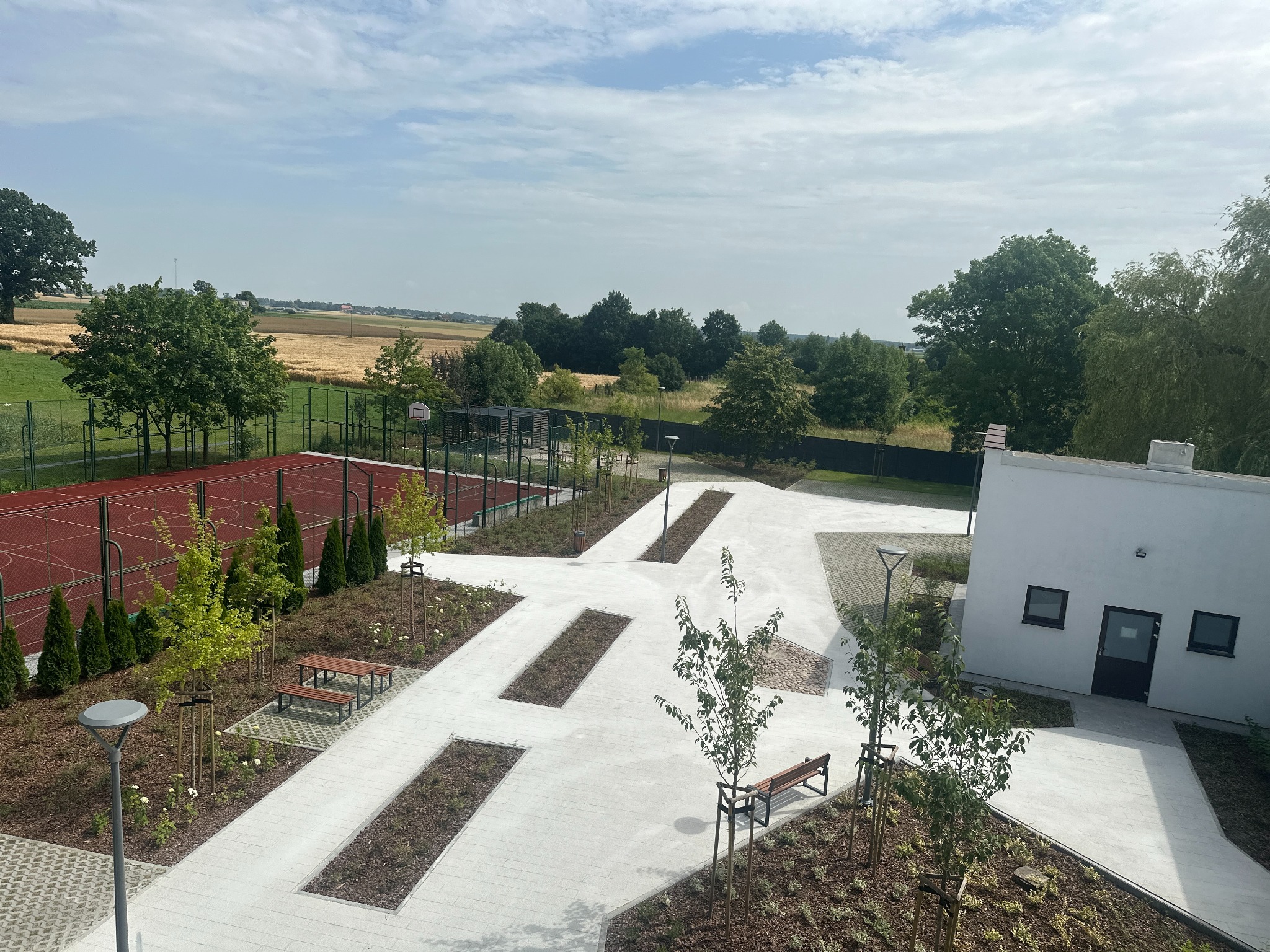 Widok z góry na nowo zagospodarowany teren z jasną kostką brukową, rabatami z młodymi drzewami i ławkami, obok boiska sportowego i budynku.