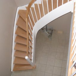 INN MARKET SP. Z O.O. - Widok z góry na spiralne schody z jasnego drewna z białą balustradą, z widoczną instalacją pod schodami i beżową ścianą w tle.