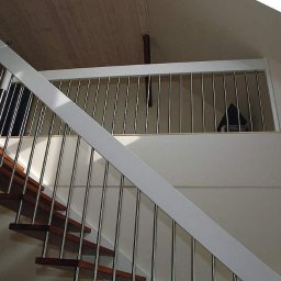 INN MARKET SP. Z O.O. - Klatka schodowa z drewnianymi stopniami i balustradą z metalowych prętów, biała konstrukcja nośna, widok z dołu na górę.