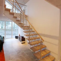 Nowoczesne schody z drewnianymi stopniami i metalową balustradą na tle jasnego wnętrza z dużym oknem.