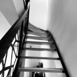 Biało-czarne ujęcie spiralnych schodów z jasnego drewna z geometryczną, metalową balustradą, widziane od dołu, z delikatnie widocznym wnętrzem pomieszczenia w tle.