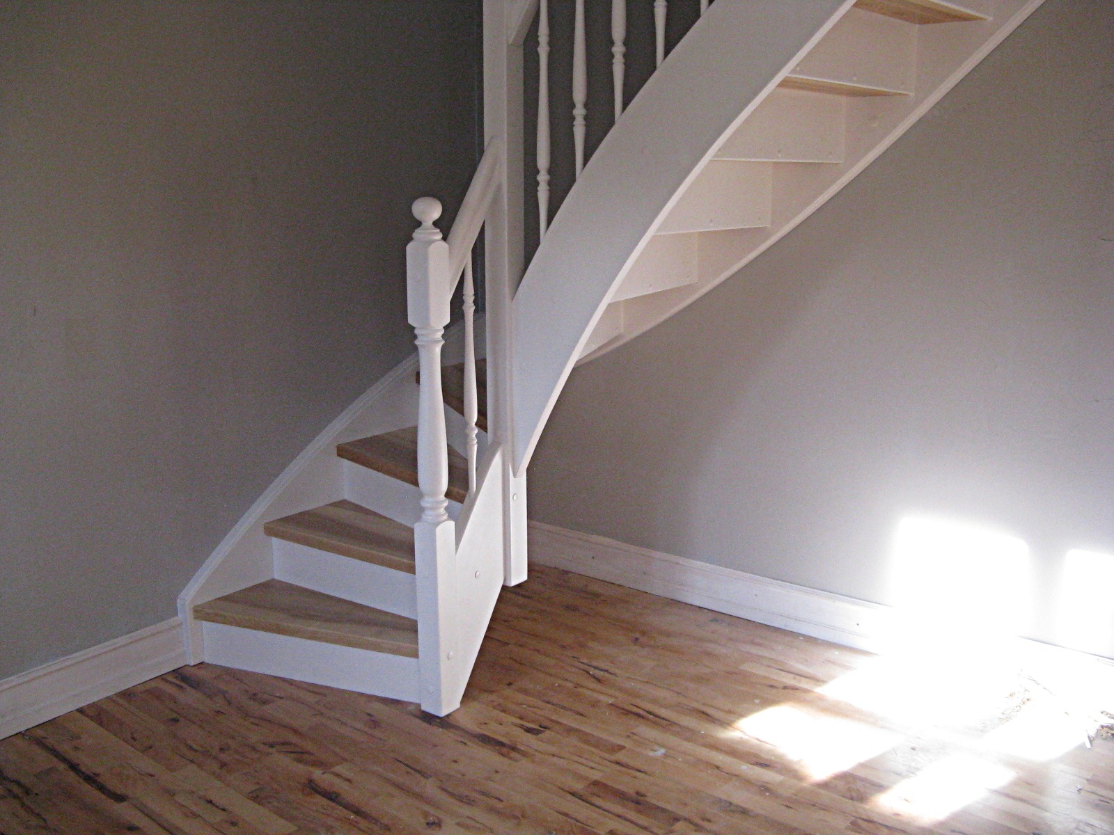 Wewnętrzne schody z drewnianymi stopniami i białą balustradą, widoczne od dołu, z fragmentem podłogi z paneli i szarą ścianą w tle.