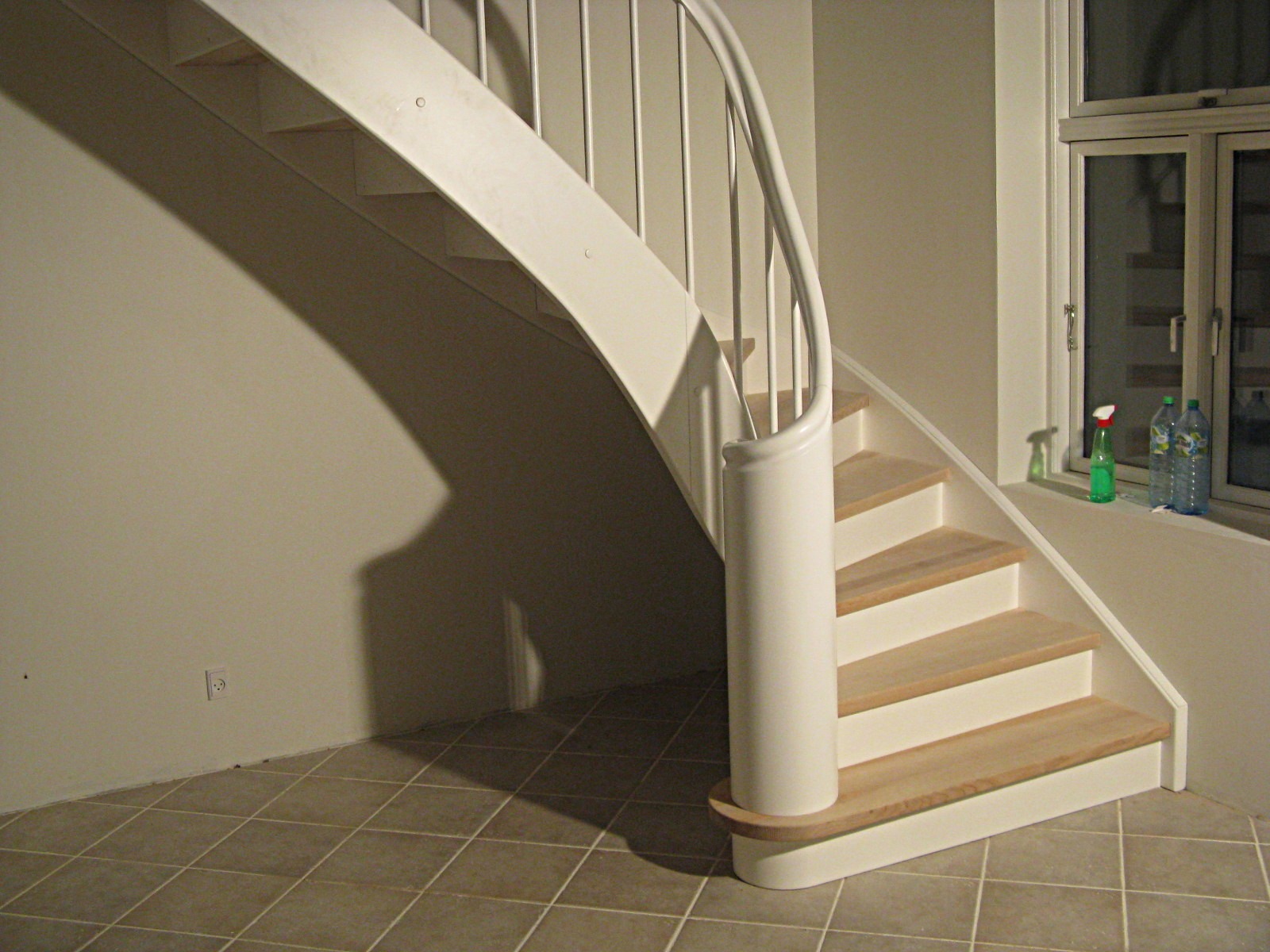 Klatka schodowa z białymi, kręconymi schodami z drewnianymi stopniami, widok z dołu, na parapecie okiennym butelki z płynami.