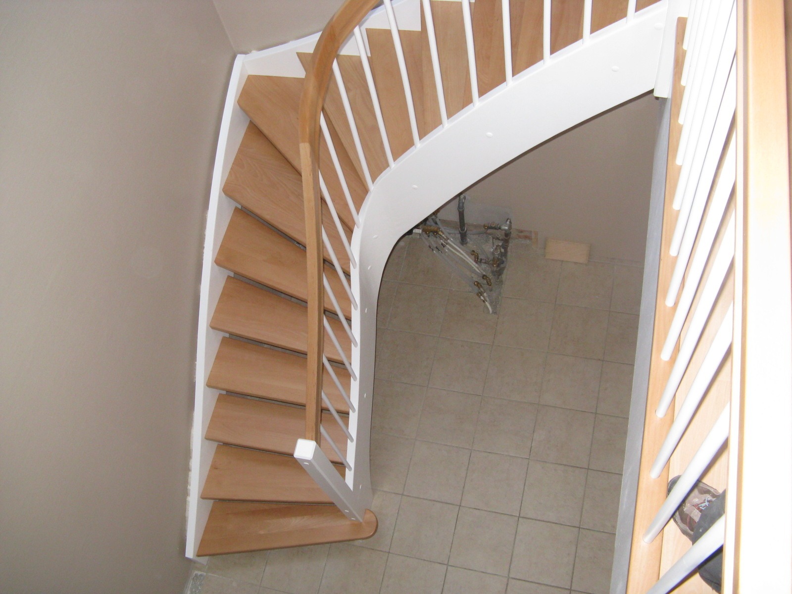 Widok z góry na spiralne schody z jasnego drewna z białą balustradą, z widoczną instalacją pod schodami i beżową ścianą w tle.