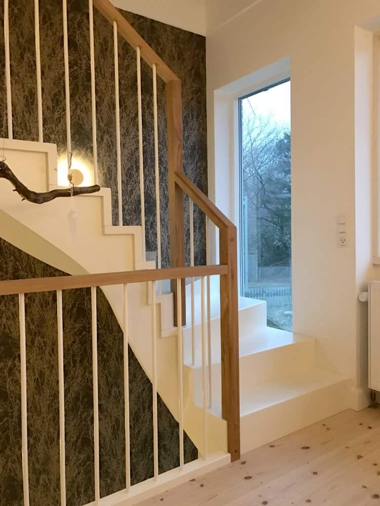 Białe schody z drewnianymi elementami i białymi balustradami w nowoczesnym wnętrzu z wzorzystą tapetą w tle i widokiem na zewnątrz przez okno.