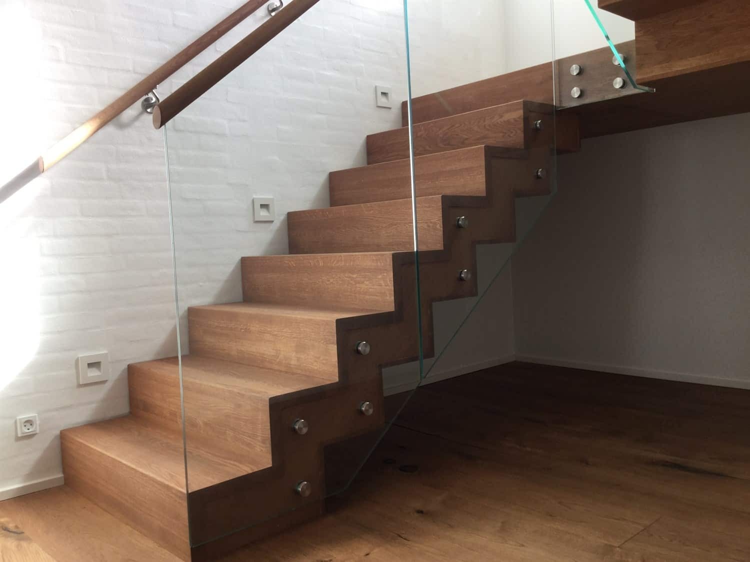 Dębowe schody z przezroczystą szklaną balustradą mocowaną metalowymi śrubami, widoczne oświetlenie punktowe na ścianie z cegły oraz drewniana podłoga.