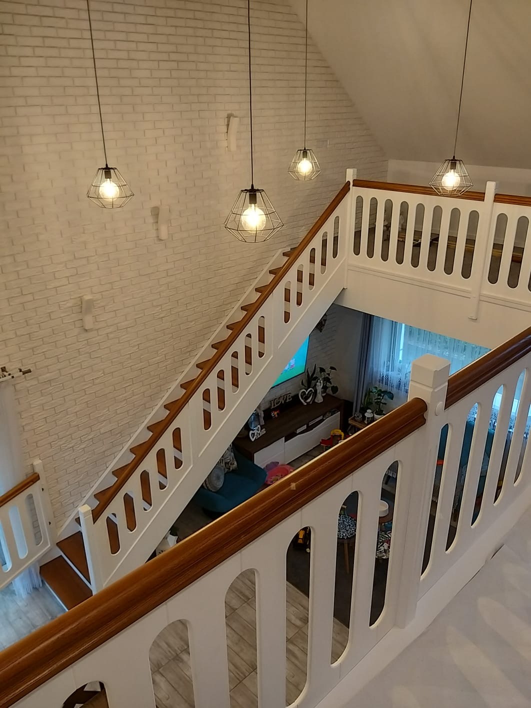 Widok z góry na nowoczesne schody z drewnianymi stopniami i białą balustradą z drewnianą poręczą, oświetlone wiszącymi lampami w stylu industrialnym, na tle białej ściany z cegły i salonu.