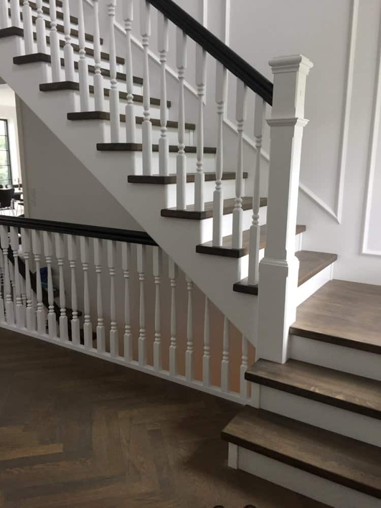 Dwukolorowe schody z drewnianymi stopniami i białymi balustradami z czarnym poręczą, na parkiecie ułożonym w jodełkę.