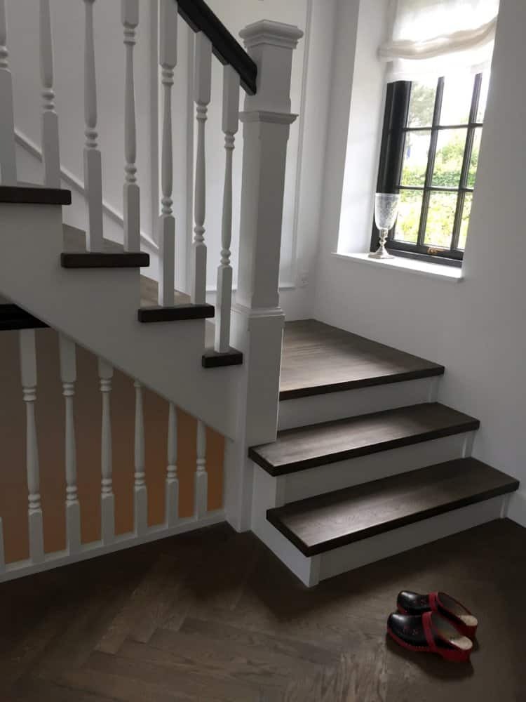 Klasyczne schody z drewnianymi stopniami o ciemnym odcieniu i białą balustradą, widoczne buty na parkiecie u dołu schodów, okno w tle.