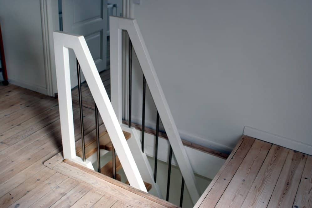 Białe schody drewniane z czarnymi metalowymi prętami balustrady, widoczne z góry, z drewnianą podłogą i białymi ścianami w tle.