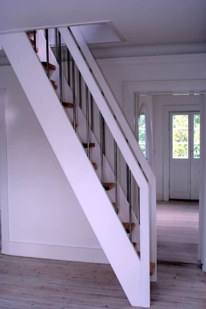 Białe schody z metalowymi balustradami w jasnym wnętrzu, widoczne drewniane stopnie i podłoga z desek.