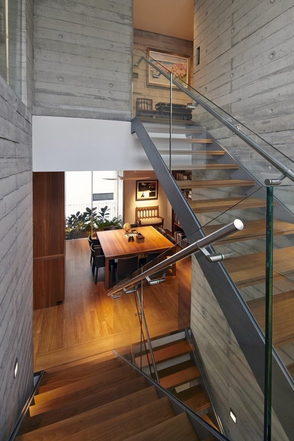 Wielopoziomowe schody z drewnianymi stopniami i szklanymi balustradami w nowoczesnym domu z betonowymi ścianami, widok na jadalnię z drewnianym stołem i krzesłami.