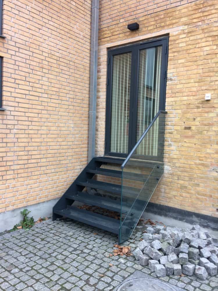 Metalowe schody zewnętrzne z szklaną balustradą i pojedynczą poręczą, prowadzące do ciemnych drzwi w ceglanym budynku, z kostką brukową i stertą kamieni u podstawy.