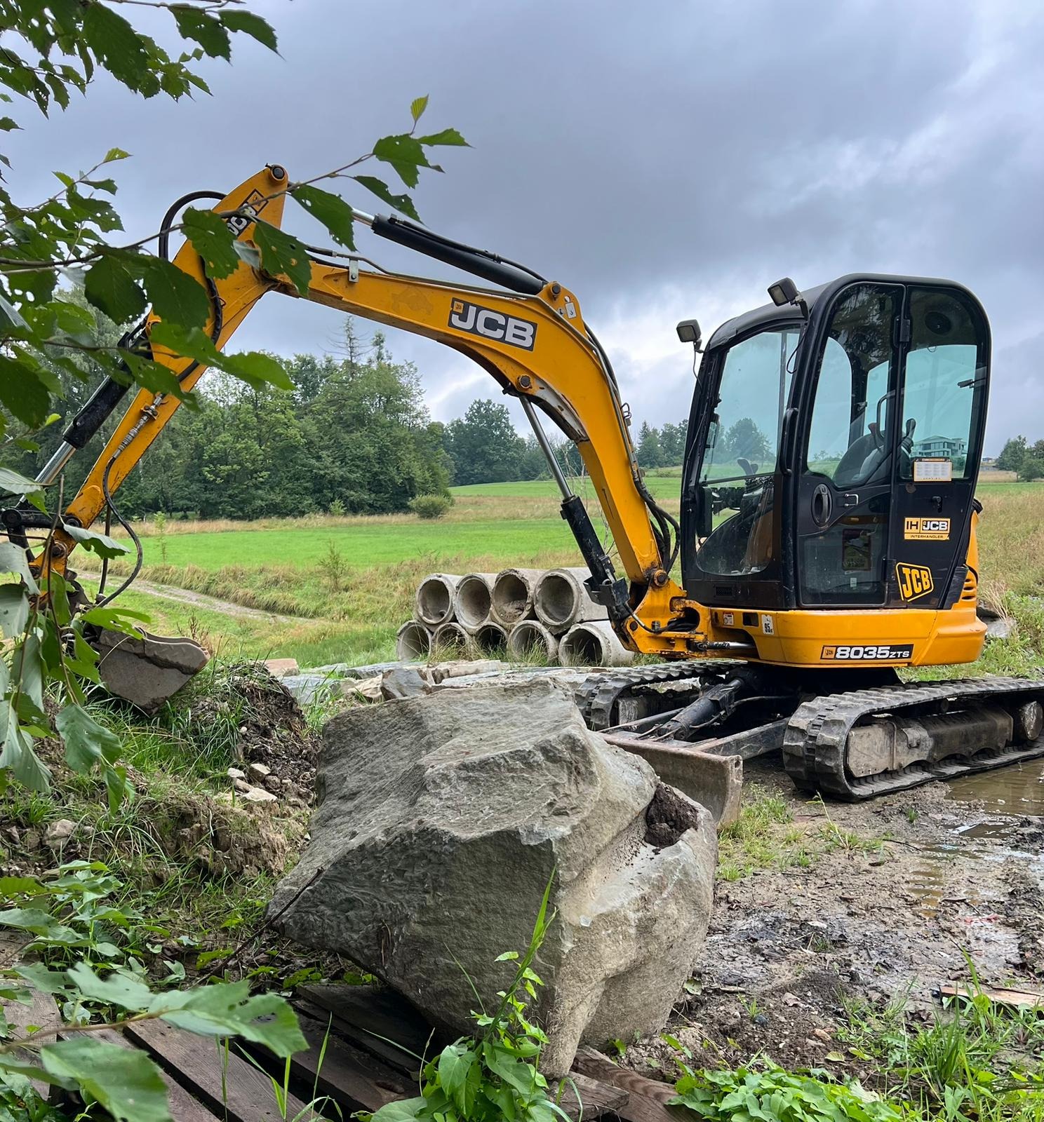 Żółta koparka JCB z łyżką obok sterty betonowych rur na tle zielonego pola i pochmurnego nieba, z dużym kamieniem na pierwszym planie i błotnistym podłożem.