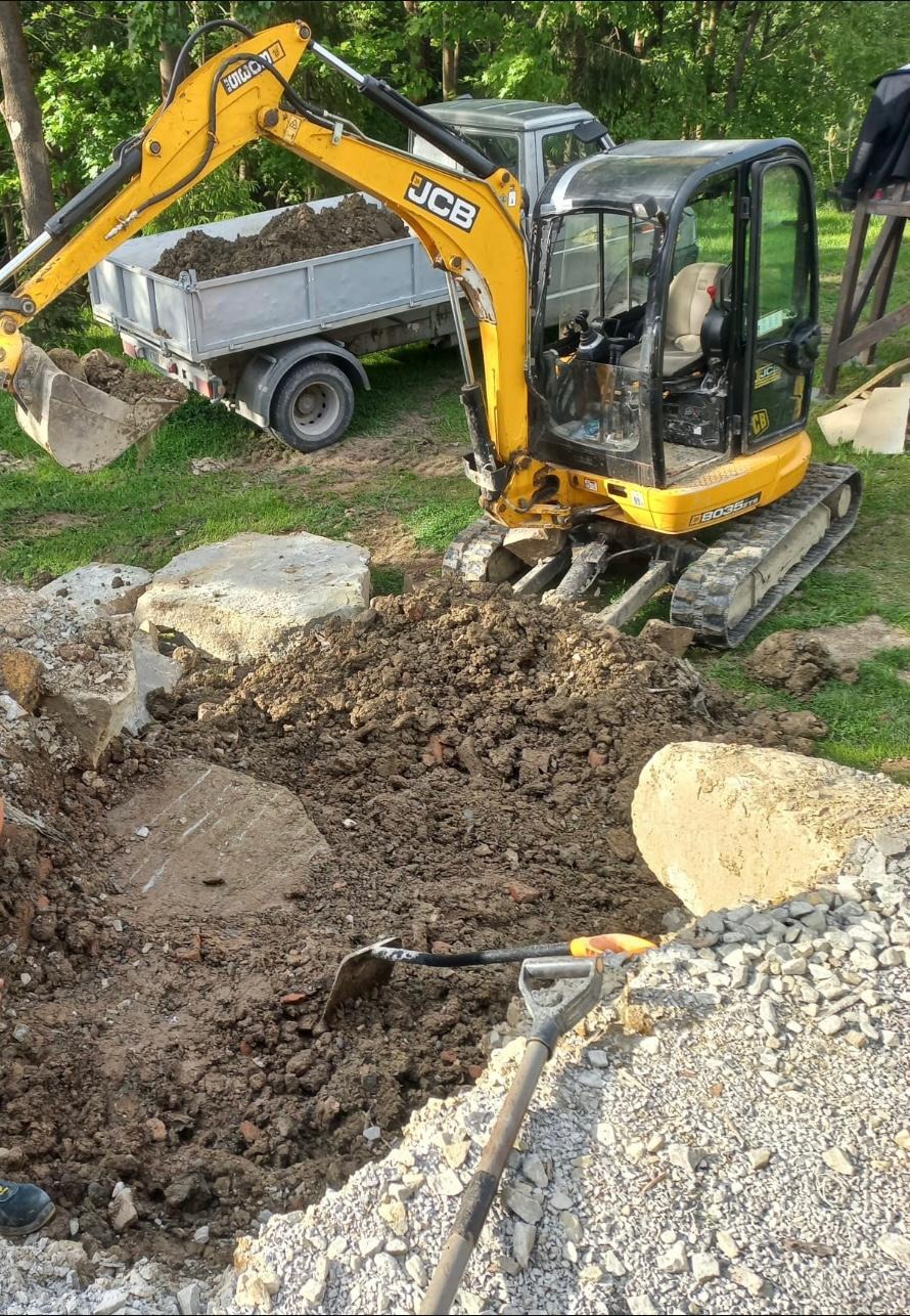 Żółta mini koparka JCB podczas prac ziemnych, łyżka nabiera ziemię do przyczepki, widoczne wykopane zagłębienie w ziemi, kamienie i dwie łopaty.