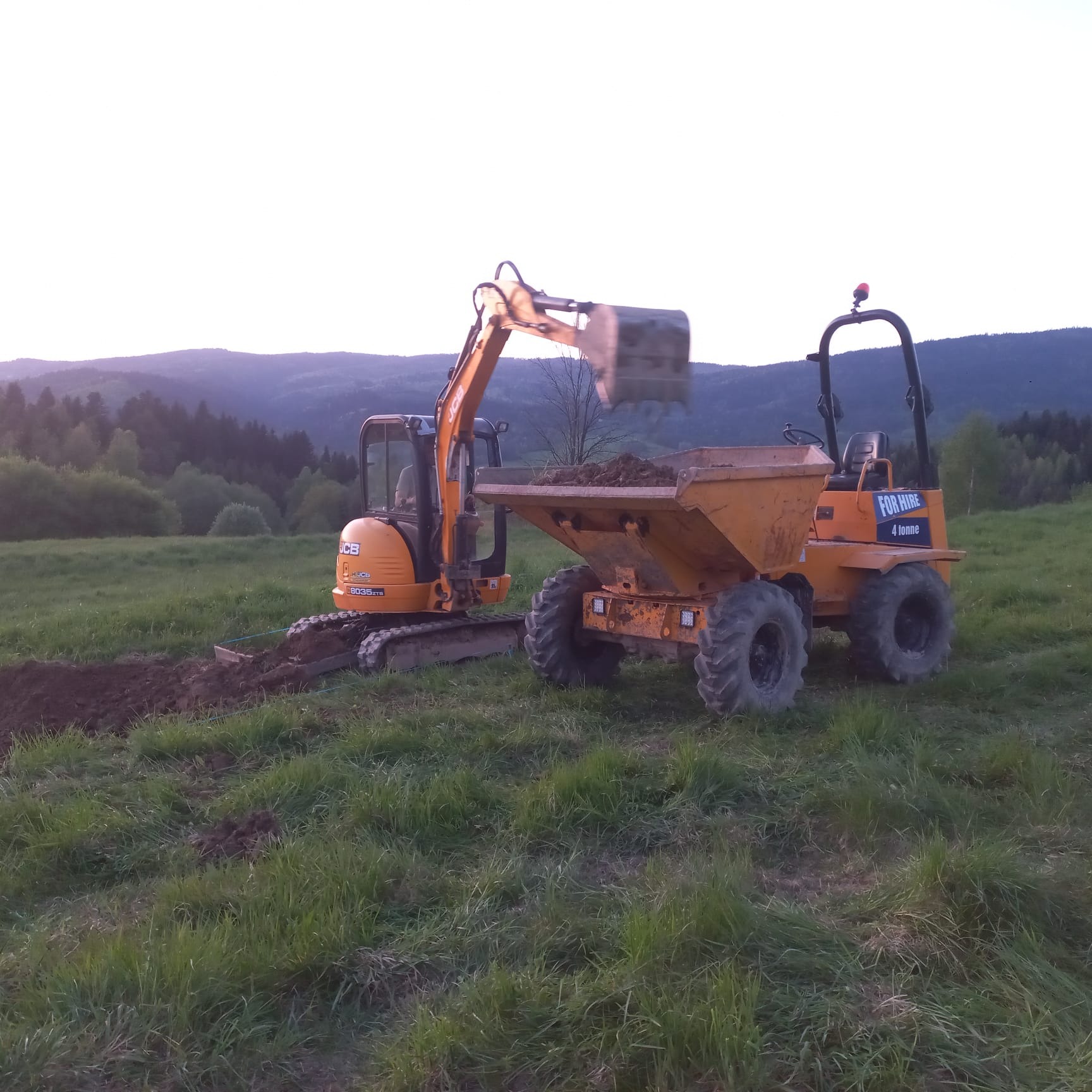 Pomarańczowa mini koparka JCB ładuje ziemię do wywrotki na tle zielonej łąki i zalesionych wzgórz w oddali.