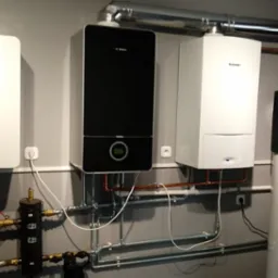 Kompletna instalacja grzewcza z trzema różnymi piecami gazowymi firm Bosch i Junkers oraz zasobnikiem ciepłej wody, widoczne rury miedziane i stalowe, szara ściana w tle.