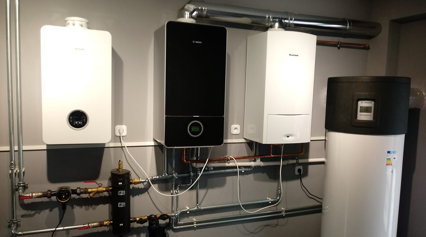 Kompletna instalacja grzewcza z trzema różnymi piecami gazowymi firm Bosch i Junkers oraz zasobnikiem ciepłej wody, widoczne rury miedziane i stalowe, szara ściana w tle.
