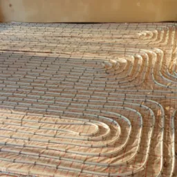 Białe rury ogrzewania podłogowego ułożone na folii z czerwoną siatką, widoczne niebieskie rury i fragment ściany w surowym stanie, ciepłe oświetlenie.