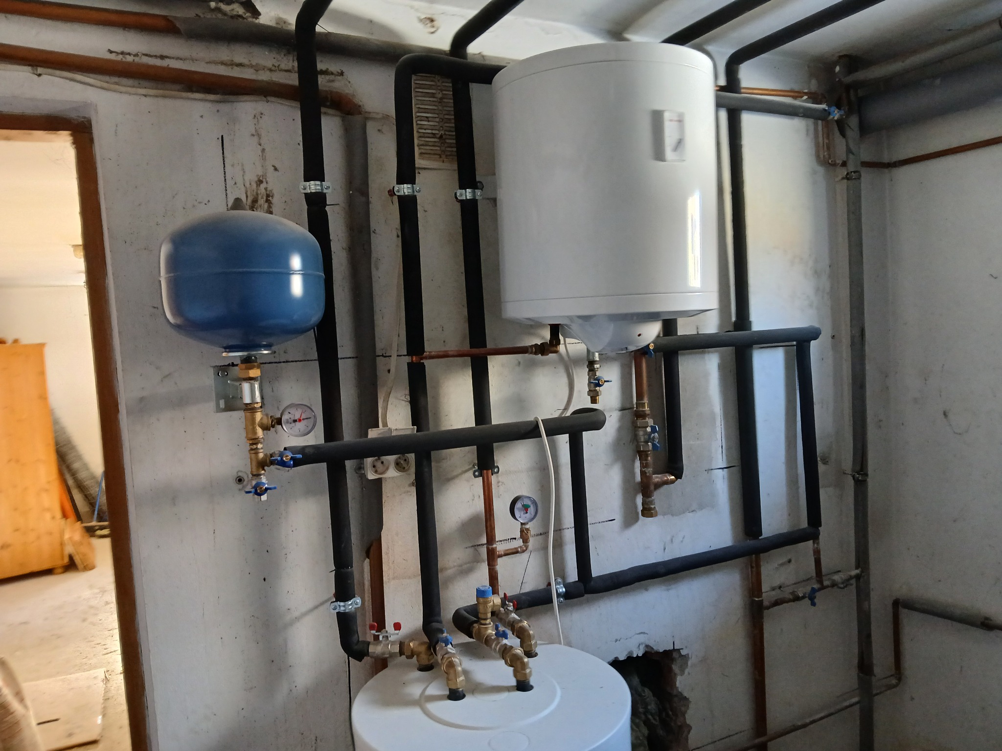 Instalacja centralnego ogrzewania z białym zasobnikiem ciepłej wody, niebieskim naczyniem przeponowym, czarnymi rurami izolowanymi i miedzianymi rurkami, zamontowana na szarej, surowej ścianie...