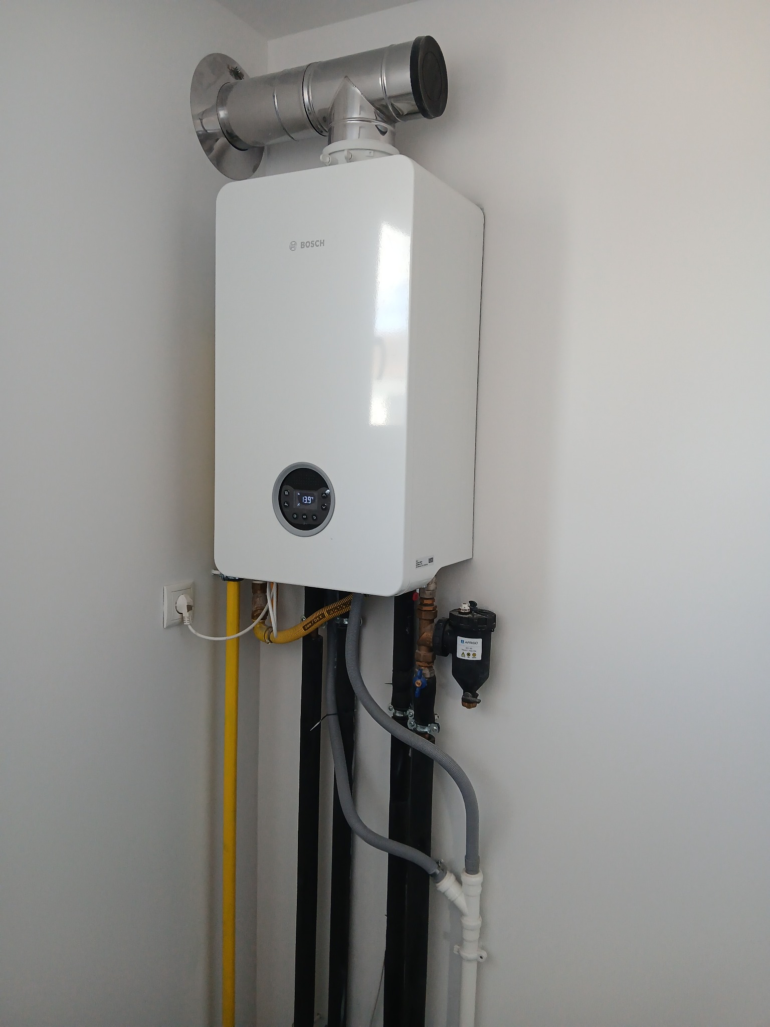 Biały, wiszący piec gazowy marki Bosch z rurą spalinową i filtrem, zamontowany na białej ścianie, z widocznymi podłączeniami gazowymi i hydraulicznymi.