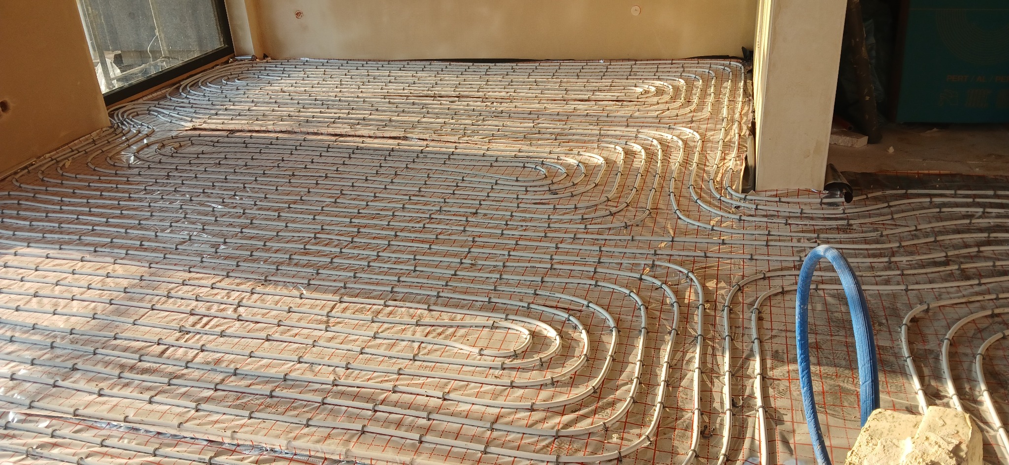 Białe rury ogrzewania podłogowego ułożone na folii z czerwoną siatką, widoczne niebieskie rury i fragment ściany w surowym stanie, ciepłe oświetlenie.