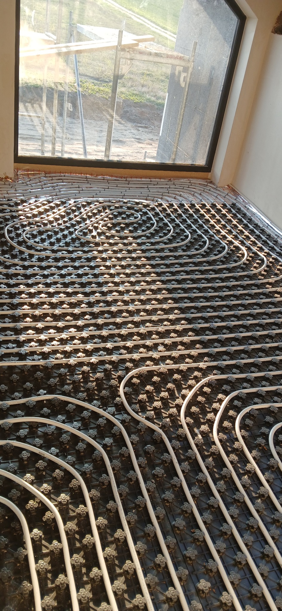Białe rury ogrzewania podłogowego ułożone spiralnie na czarnej macie, z widokiem na okno i plac budowy w tle. Duże nasłonecznienie.