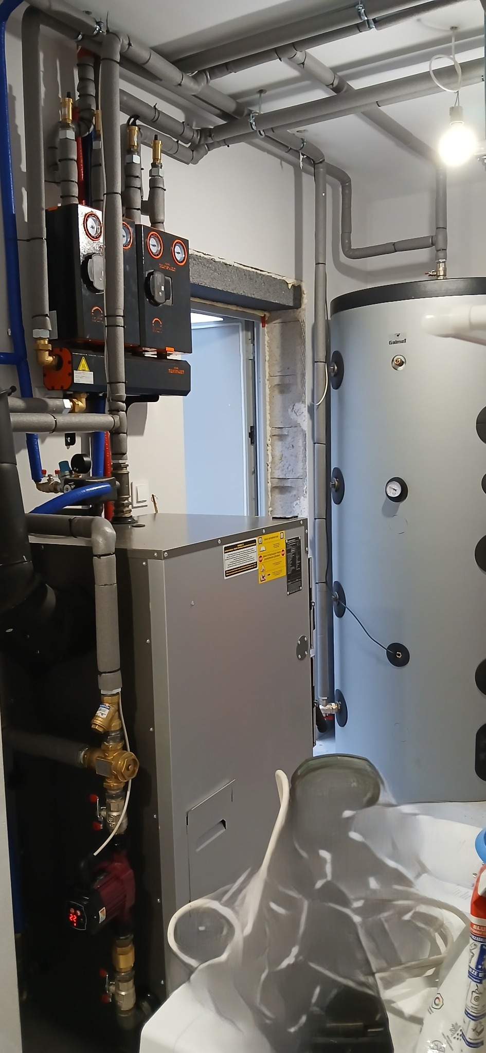 Złożona instalacja grzewcza z kotłem, zasobnikiem i rurami w szarych izolacjach. Widoczne elementy sterujące i zabezpieczające. Nowoczesne rozwiązanie w kotłowni.