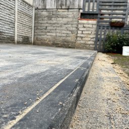 AS-BUD Usługi brukarskie - Ułożona, szara kostka brukowa z czarnym obrzeżem, zasypana piaskiem, widok z niskiej perspektywy na tle zabudowań gospodarczych.