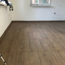 AS-BUD Usługi brukarskie - Wnętrze w trakcie prac wykończeniowych; ułożona podłoga z płytek imitujących drewno, poziomica laserowa na statywie, ściany częściowo przygotowane do malowania, widoczne puszki elektryczne.