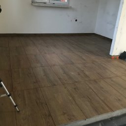 AS-BUD Usługi brukarskie - Układanie podłogi z płytek imitujących drewno w jasnym pomieszczeniu, widoczny laser krzyżowy na statywie.
