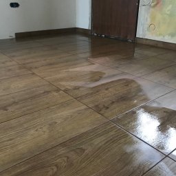 AS-BUD Usługi brukarskie - Świeżo ułożona, błyszcząca podłoga z płytek imitujących drewno. Widoczny kubeł z zaprawą i poziomica w rogu pomieszczenia. Częściowo pomalowana ściana w tle.