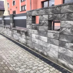 Nowoczesny mur z szarej kostki betonowej o nieregularnym wzorze, z otworami w górnej części, przylegający do chodnika z kostki brukowej i ogrodzenia z szarymi panelami.
