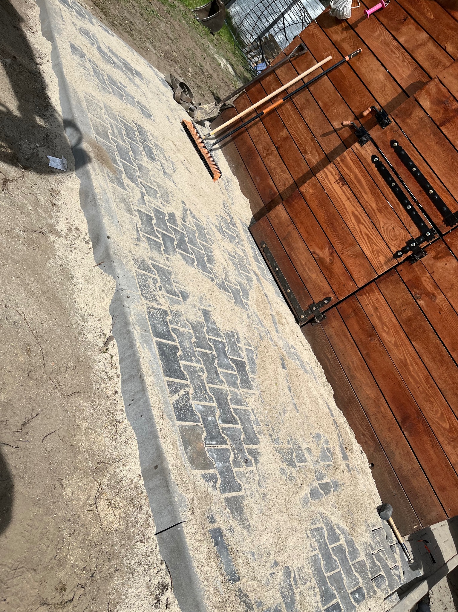 Ukończona część ścieżki z kostki brukowej, zasypana piaskiem, z widocznymi narzędziami (szczotka, młotek) i drewnianą bramą w tle.