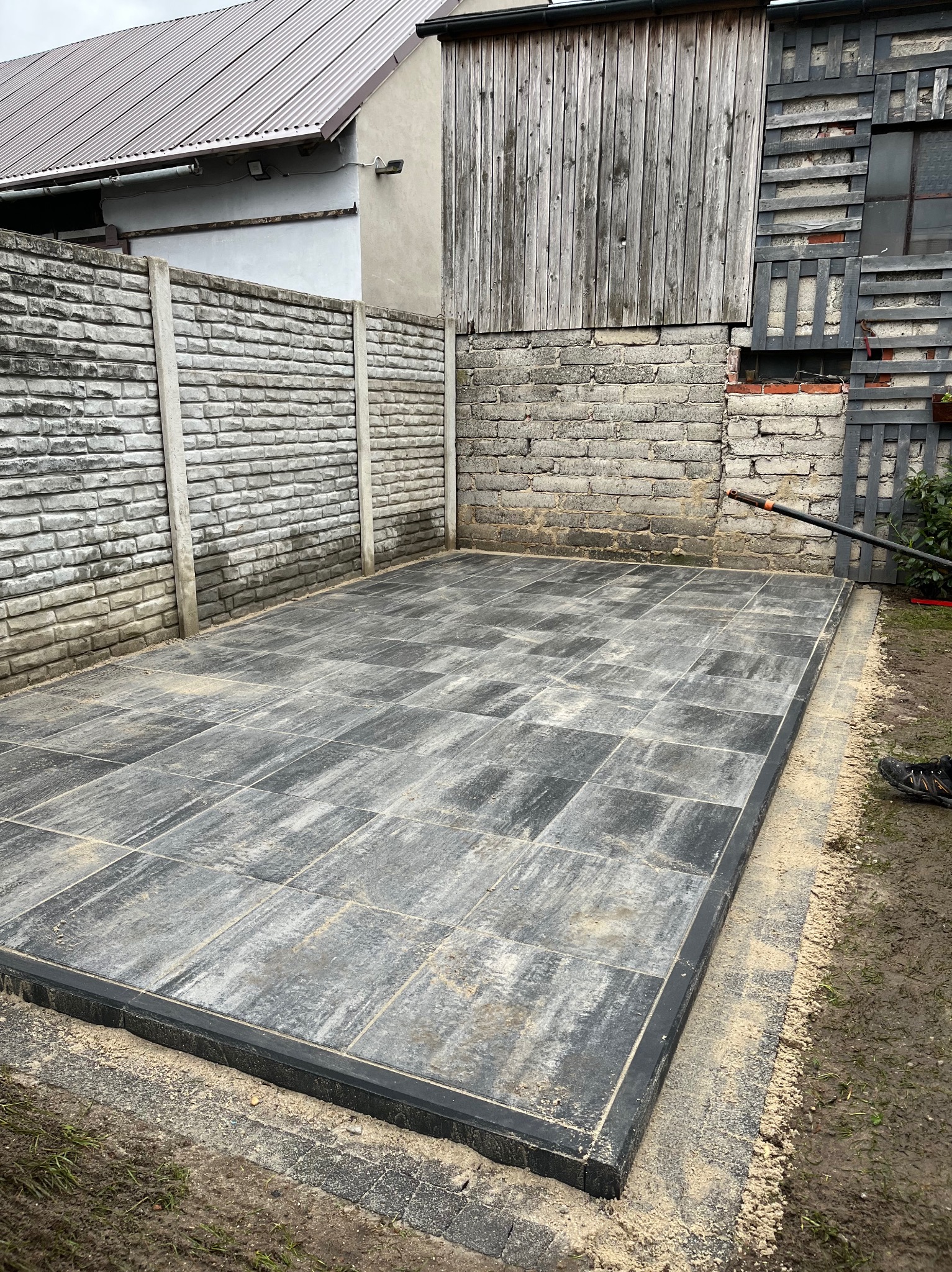 Ułożona szara kostka brukowa tworząca patio przy ceglanym i betonowym ogrodzeniu, widoczne ślady piasku na powierzchni i czarne obrzeże, w tle budynek gospodarczy z drewnianą ścianą.