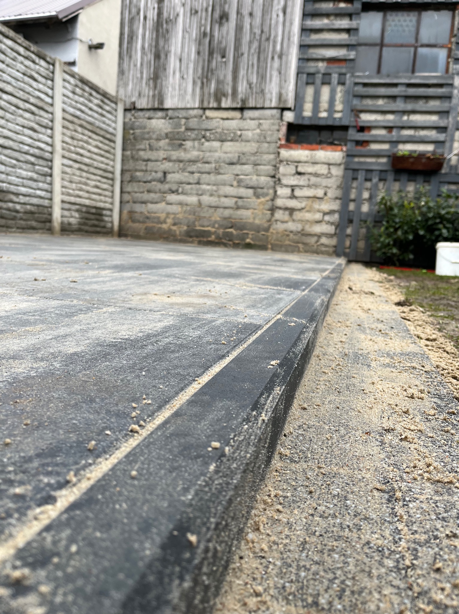 Ułożona, szara kostka brukowa z czarnym obrzeżem, zasypana piaskiem, widok z niskiej perspektywy na tle zabudowań gospodarczych.