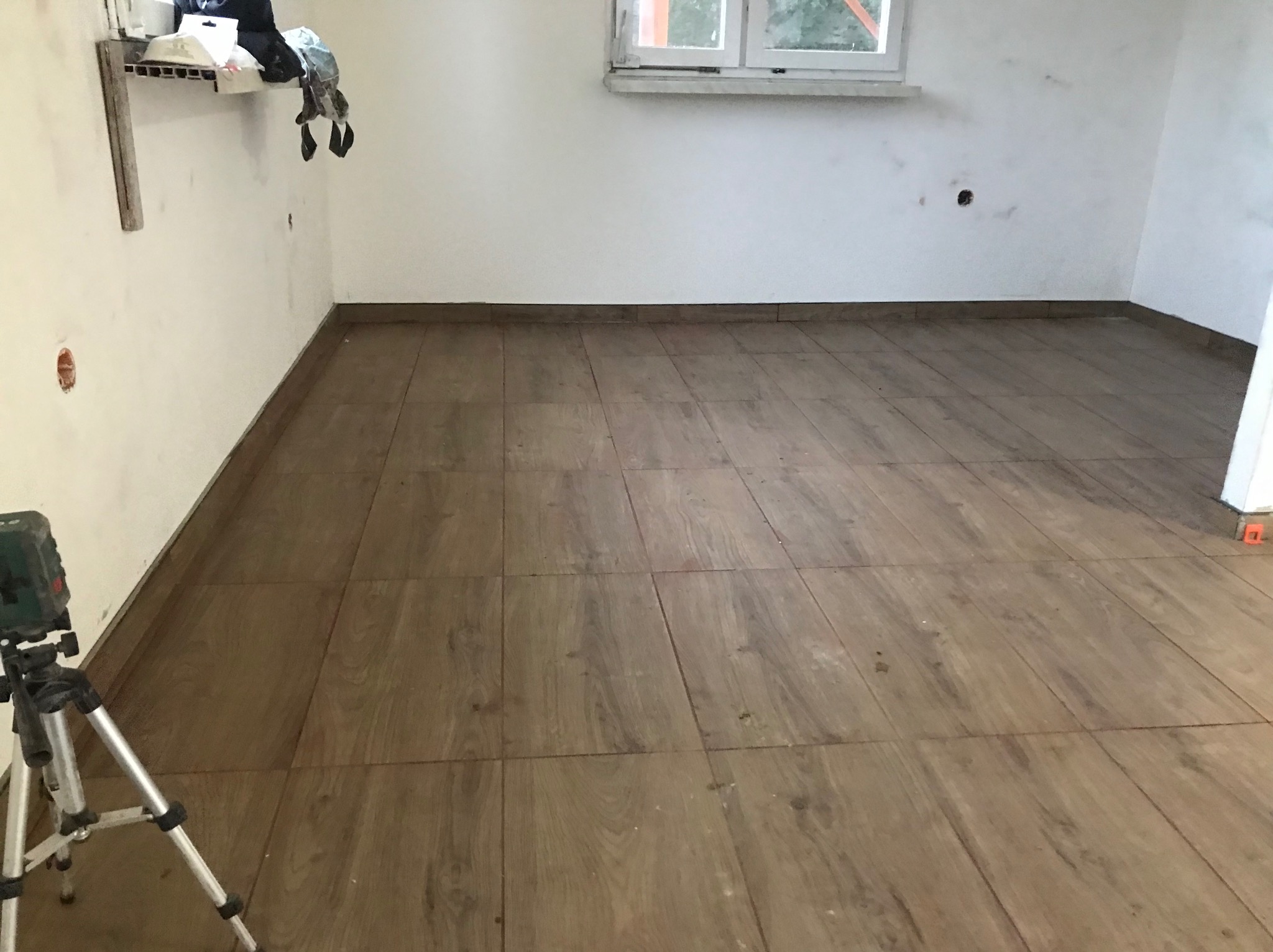 Wnętrze w trakcie prac wykończeniowych; ułożona podłoga z płytek imitujących drewno, poziomica laserowa na statywie, ściany częściowo przygotowane do malowania, widoczne puszki elektryczne.