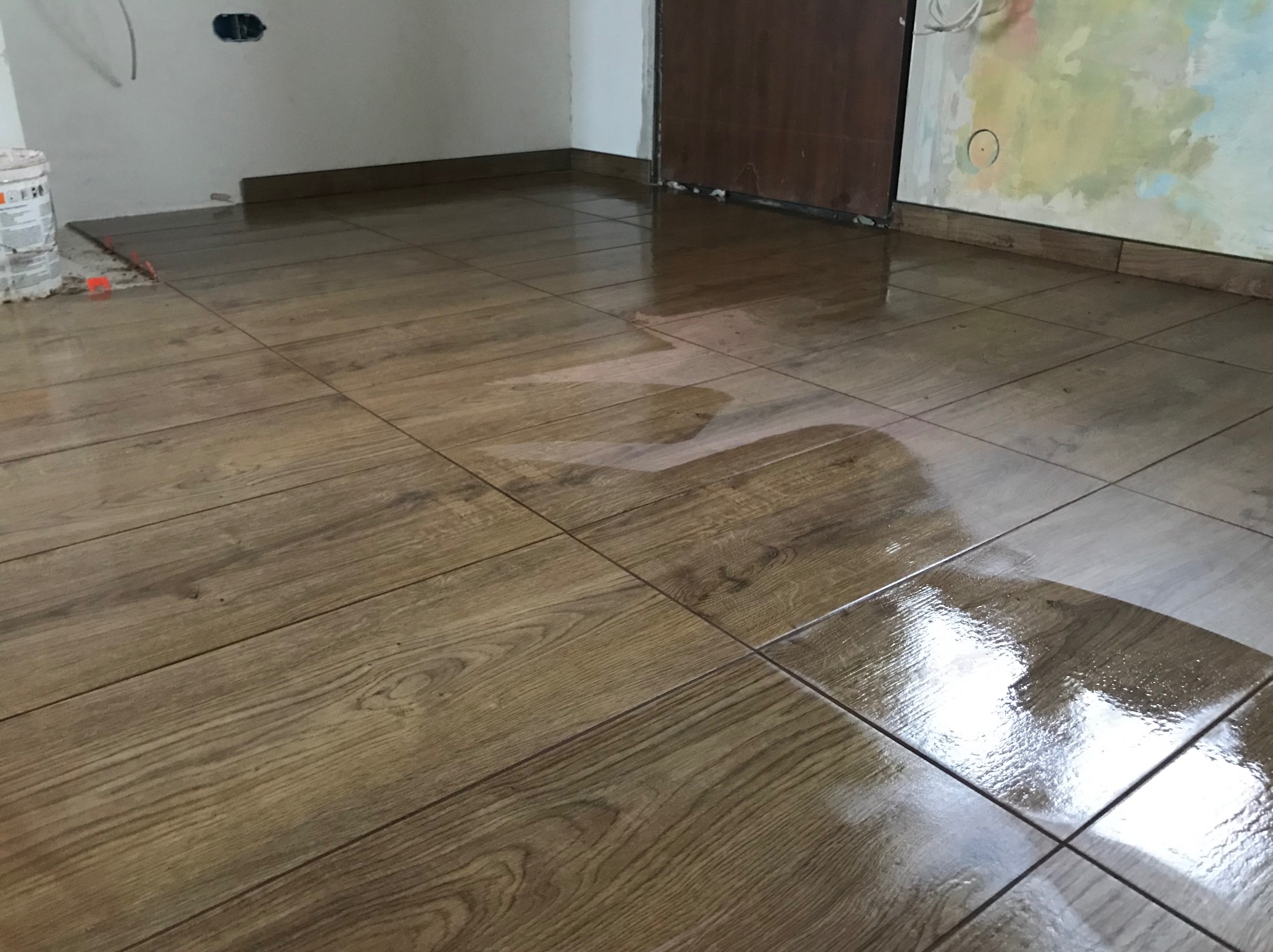 Świeżo ułożona, błyszcząca podłoga z płytek imitujących drewno. Widoczny kubeł z zaprawą i poziomica w rogu pomieszczenia. Częściowo pomalowana ściana w tle.